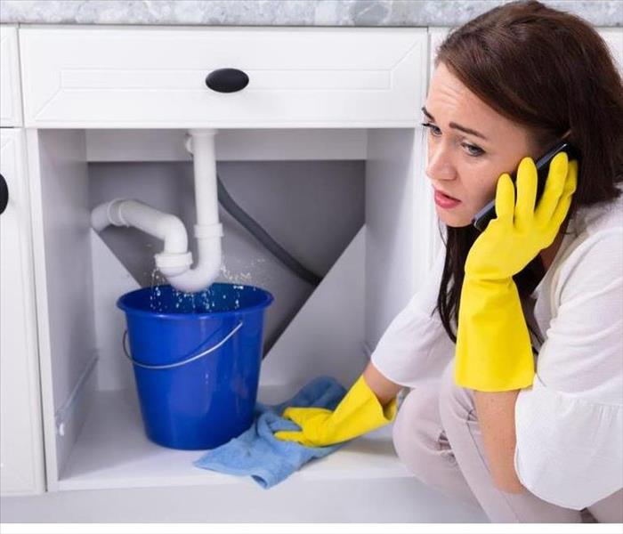 A woman on the phone trying to stop a leaking faucet under a sink 